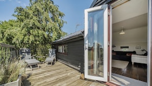 A wooden deck with outdoor furniture, a sliding glass door leading to a room with a bed and a sofa, and a large tree in the background. - Hayloft-Sleeps 3-with Enclosed terrace (Dereham)