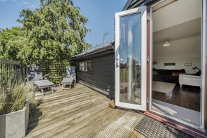 A wooden deck with outdoor furniture, a sliding glass door leading to a room with a bed and a sofa, and a large tree in the background. - Hayloft-Sleeps 3-with Enclosed terrace (Dereham)