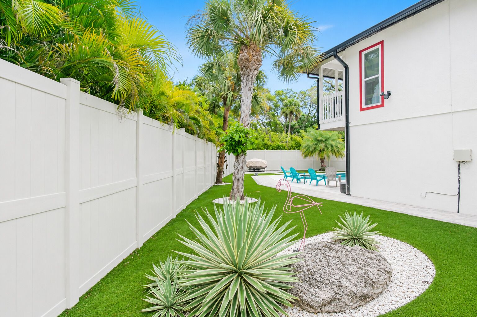 A backyard with artificial grass, a white fence, a palm tree, and outdoor furniture.