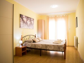 Um quarto com uma cama, mesa de cabeceira, abajur, quadro na parede e janela com cortinas.