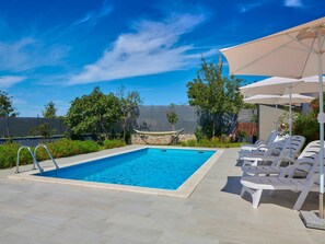 Piscina retangular com um deck ladrilhado, rodeada por cadeiras de descanso brancas e uma área coberta com guarda-sóis.