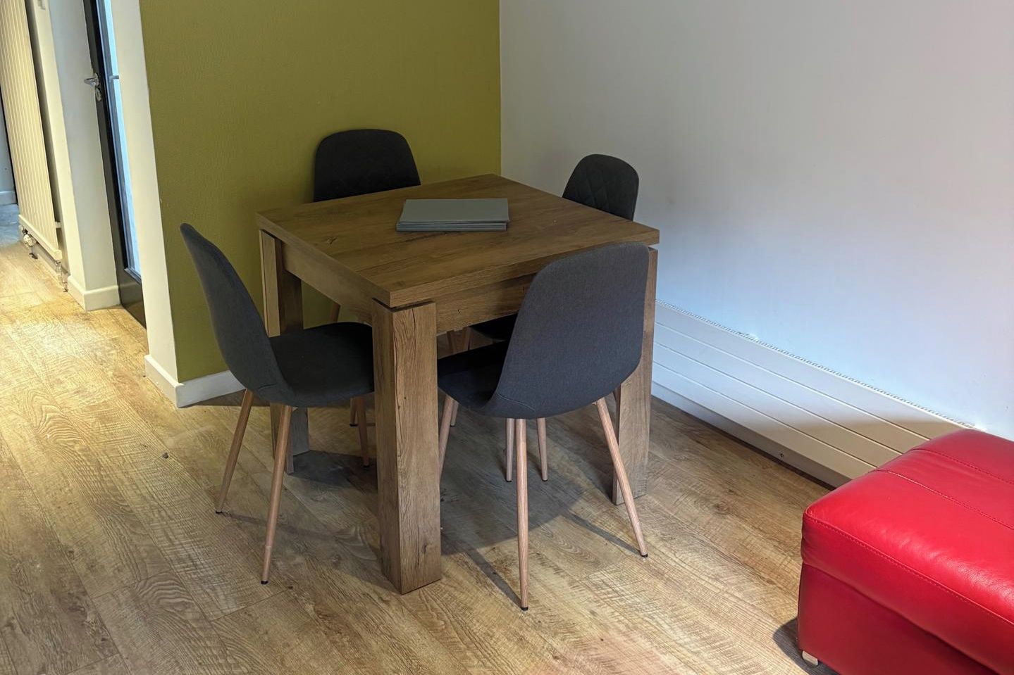 A wooden dining table with four chairs, a red ottoman, and a window with blinds.