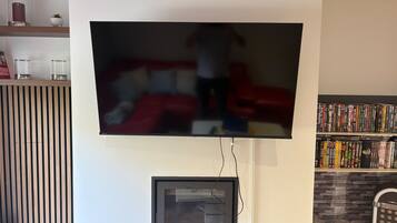 A flat-screen TV mounted on a wall, a framed picture below it, a small shelf with decorative items, and a bookshelf with DVDs.