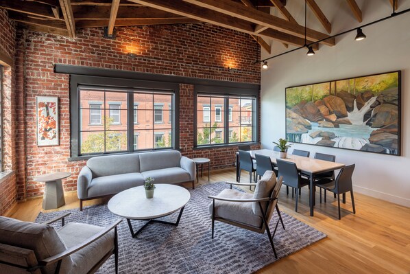 A modern interior with exposed brick walls, large windows, a gray sofa, round coffee table, and a dining area with chairs and a table.