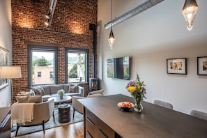 Un salon moderne avec un mur en briques, de grandes fenêtres, une cheminée et un coin repas comprenant une table et des chaises.