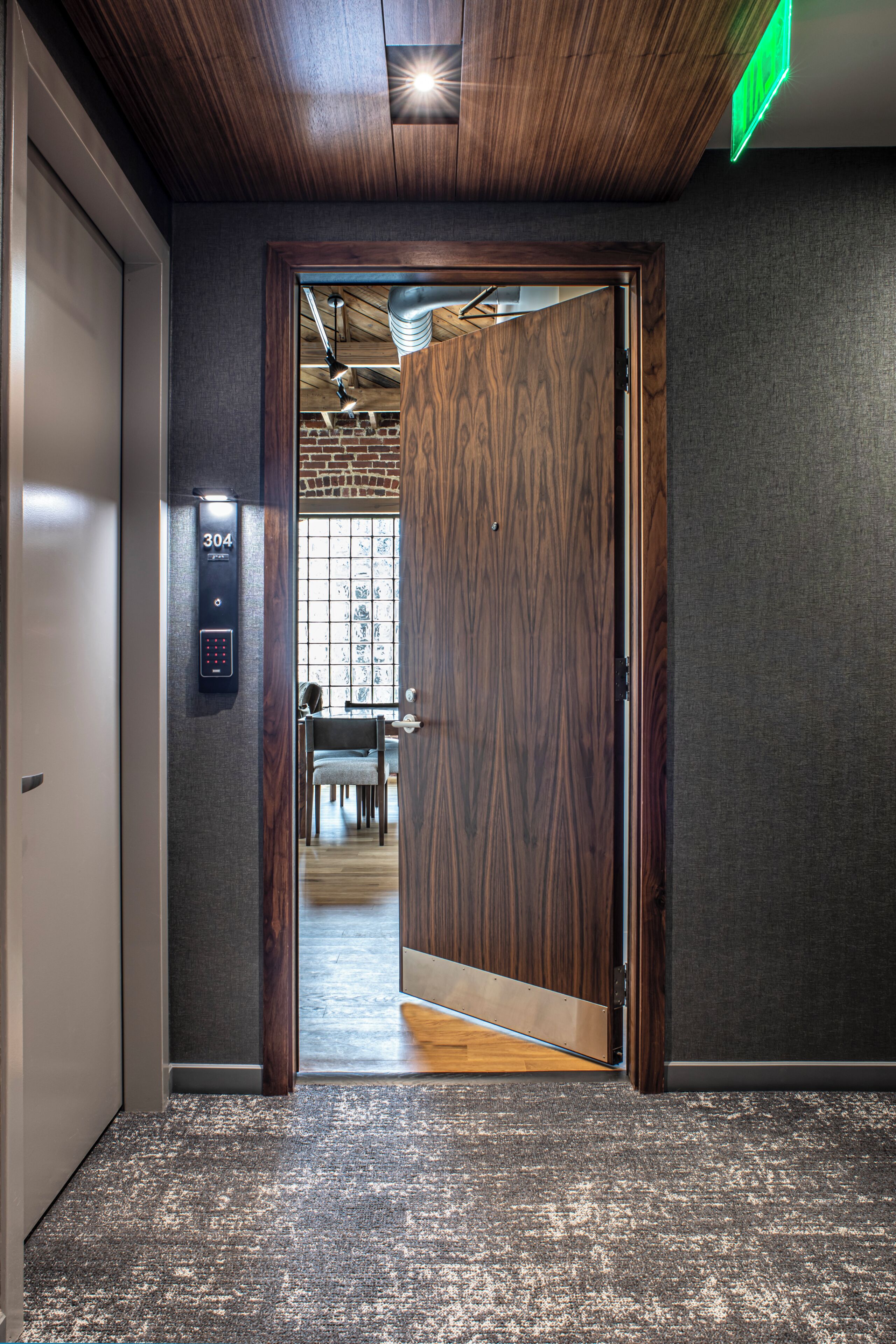 Un couloir intérieur avec une porte en bois, un panneau de commande numérique pour l’ascenseur, et une vue sur un coin repas avec un mur en briques et une fenêtre.