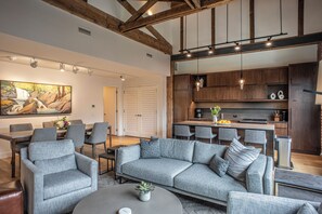 Un salon moderne avec un canapé gris, des fauteuils gris et un coin repas comprenant une table et des chaises. On y trouve également une cuisine avec des meubles en bois et une table à manger.