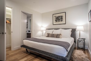 Een verzorgde slaapkamer met een groot bed, bedlampen, een lijst met een kunstwerk aan de muur en een kast met hangende kleding.