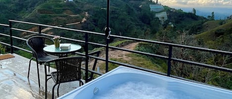 A hot tub on a wooden deck with a table and chairs, an umbrella, and a view of a hilly landscape.