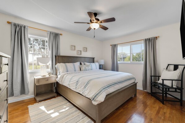 Un dormitorio con cama, mesitas de noche, una silla, ventilador de techo y ventana con cortinas.