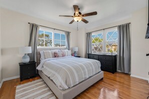 A bedroom with a bed, two nightstands, a ceiling fan, and large windows with curtains.