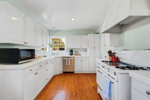 Una cocina con gabinetes blancos, lavavajillas de acero inoxidable, microondas negro, hervidor rojo y estufa blanca.