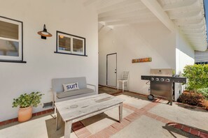 Un patio exterior con una pared blanca, un sofá gris con un cojín que dice 'Surf', una mesa de madera, una parrilla y una planta en maceta.