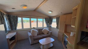 A compact RV interior with a sofa, a small table, a kitchenette, and large windows offering a view of the outside. - Luxury Caravan situated on Holistic wellbeing site (Mablethorpe)