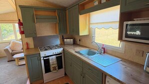 A compact kitchen with a stove, sink, and microwave, wooden cabinets, and a window providing natural light. - Luxury Caravan situated on Holistic wellbeing site (Mablethorpe)