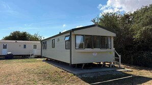 A mobile home with a covered porch and a small deck, situated in a grassy area with trees in the background. - Luxury Caravan situated on Holistic wellbeing site (Mablethorpe)