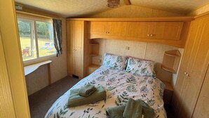 A bedroom with a bed, wooden cabinets, a window with curtains, and a view of the outside. - Luxury Caravan situated on Holistic wellbeing site (Mablethorpe)