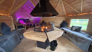 A cozy cabin interior with a central wood-burning stove, seating areas, and a window providing a view of the outdoors.