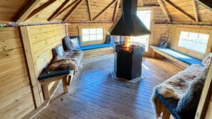 A cozy cabin interior with a wooden floor, a central fireplace, and a bench with a fur throw.