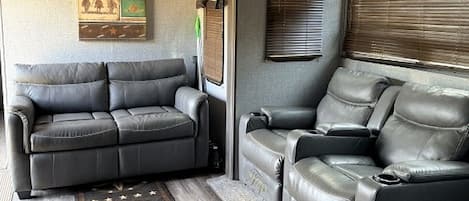 A compact RV interior with a sofa, two recliners, a ceiling fan, and a patterned rug.