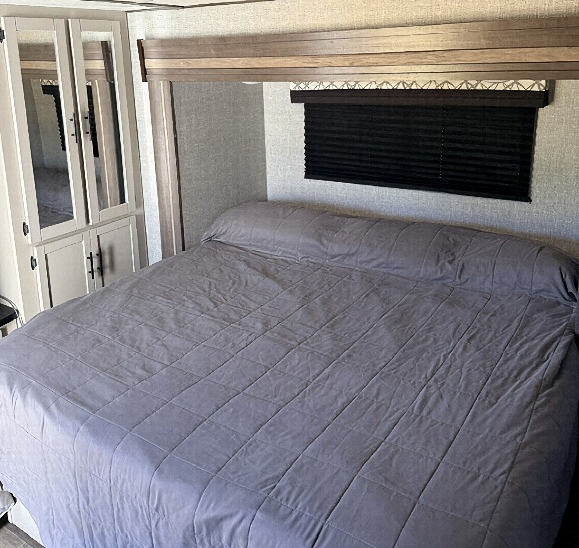 A bed with a grey quilt in a room with a wooden headboard and a window with blinds.