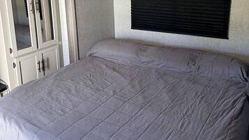 A bed with a grey quilt in a room with a wooden headboard and a window with blinds.