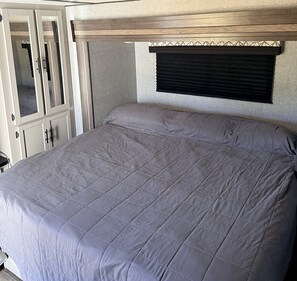 A bed with a grey quilt in a room with a wooden headboard and a window with blinds.