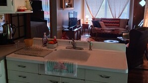 A kitchen with a central island, a dining area with a sofa and a TV, and a wooden ceiling with pendant lights.
