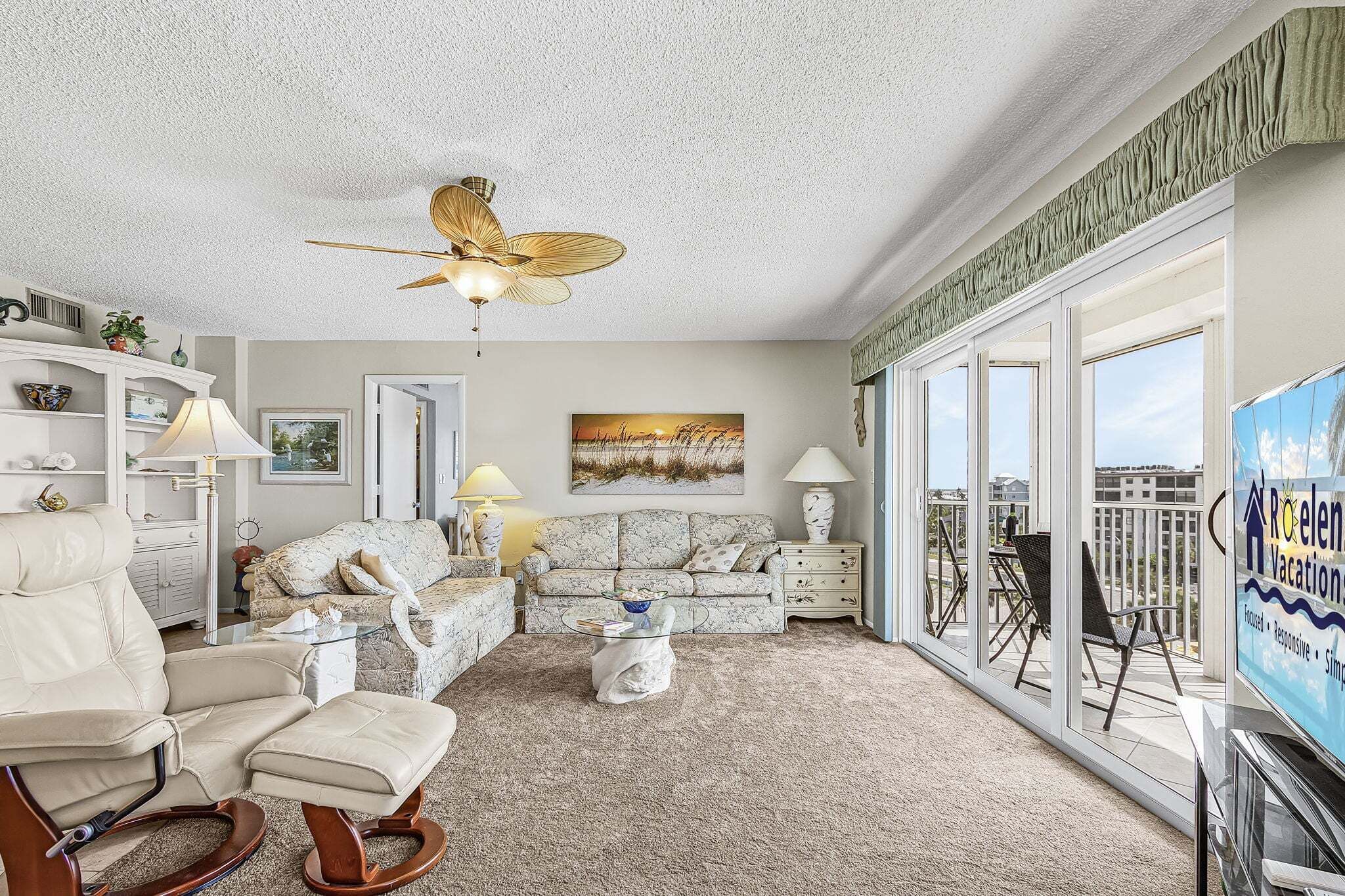 A spacious living room with a ceiling fan, a sofa, a rocking chair, a glass coffee table, and a balcony with outdoor furniture.