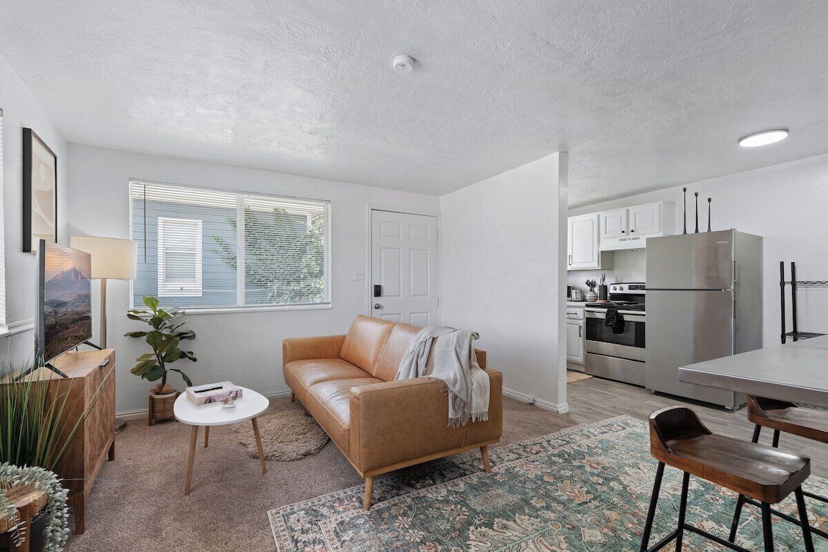 Ett kompakt bostadsutrymme med en soffa, ett litet bord, en barstol, ett köksområde med vitvaror och ett fönster med persienner.