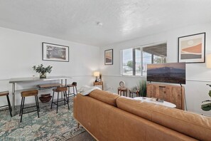 A living room with a brown leather sofa, a dining table with bar stools, and a television on a stand. - 1BR Cozy SLC Retreat Near Downtown & Trails (Salt Lake City)