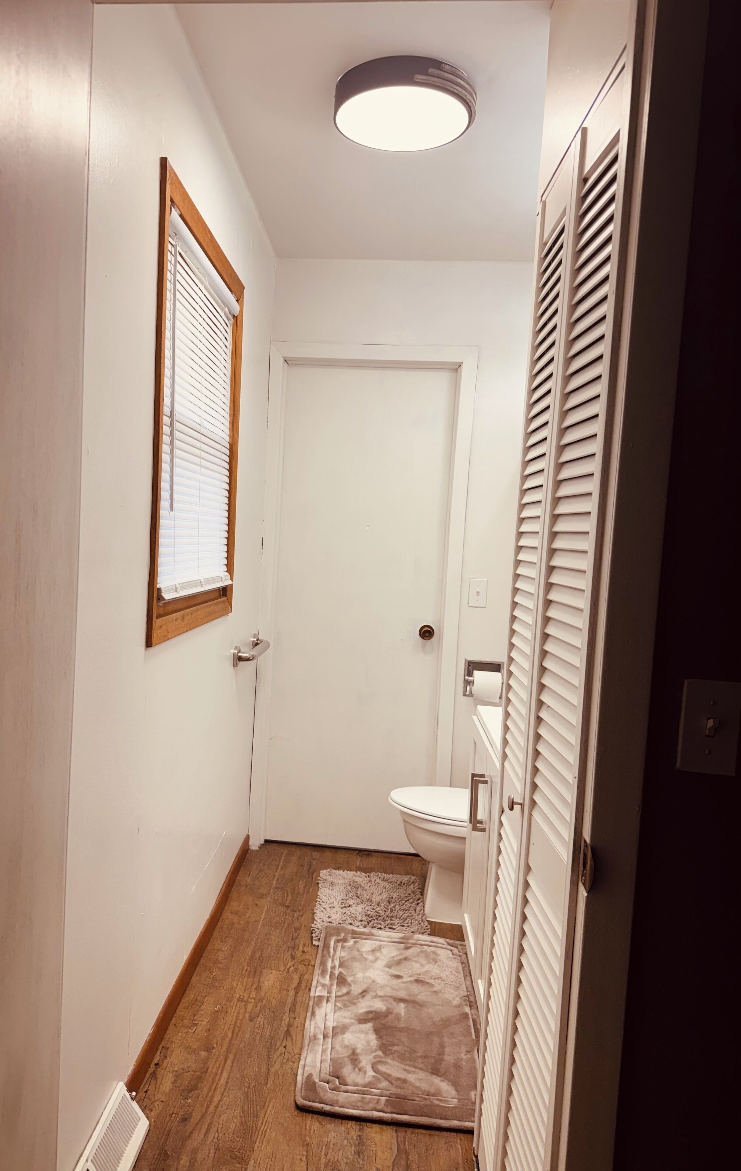 A narrow hallway with a white door, a window with blinds, a toilet, and a radiator.