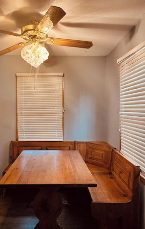 A dining area with a wooden table and bench seating, a ceiling fan, and two windows with blinds.