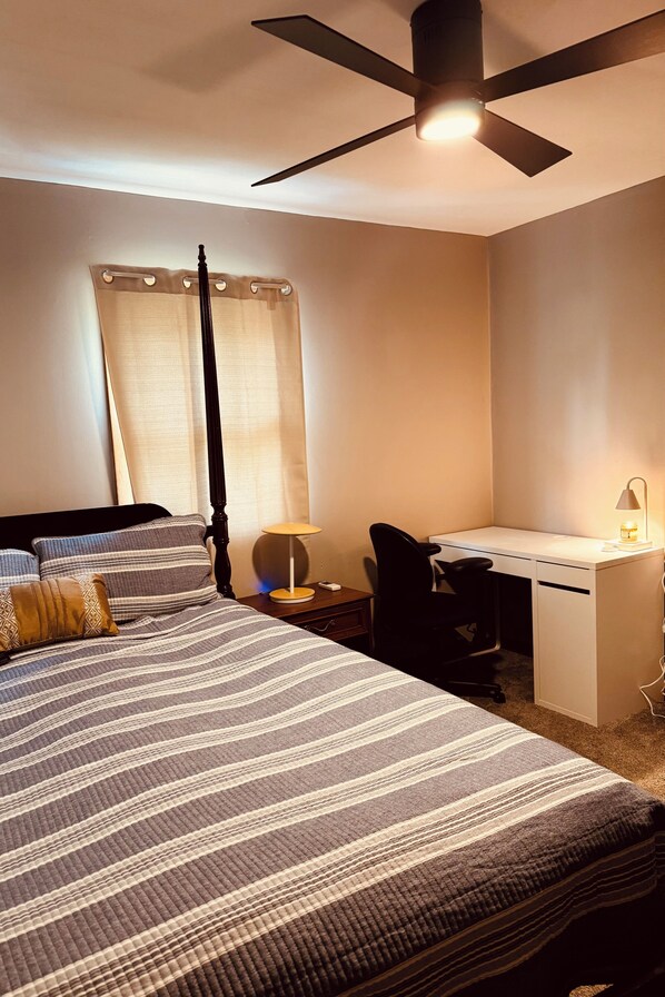 A hotel room with a bed, a desk, a chair, a ceiling fan, and a window with curtains.
