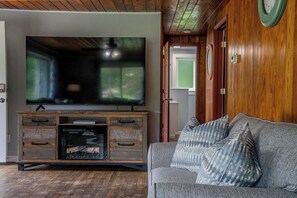 Ett vardagsrum med en platt-TV placerad på ett träskåp, en grå soffa med mönstrade kuddar och en trädörr med en rund spegel.