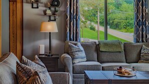 Un salon chaleureux avec un canapé, un fauteuil et une table basse. On y trouve une lampe, une étagère avec des objets décoratifs et, par une grande fenêtre, une vue sur un paysage verdoyant.