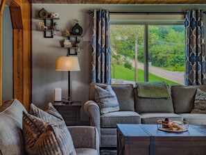 Ein gemütliches Wohnzimmer mit Sofa, Ohrensessel und Couchtisch. Eine Lampe, ein Regal mit Dekoration und Blick auf eine grüne Landschaft durch ein großes Fenster.
