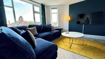 A modern living room with a blue sofa set, a round coffee table, and a flat-screen TV mounted on the wall.