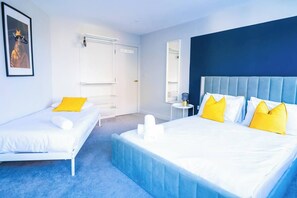Quarto de hotel com duas camas de solteiro, uma cama grande, uma parede azul decorativa, uma porta branca e um quadro na parede.