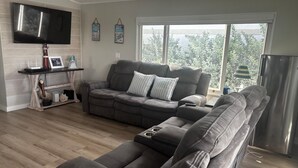 A living room with a grey sectional sofa, a flat-screen TV mounted on the wall, and a large window providing a view of greenery outside. - 2-bedroom modular home in wonderful Long Key with beautiful ocean views/sunsets (Layton)