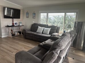 Ein Wohnzimmer mit einem grauen Ecksofa, einem flachbildschirmfernseher an der Wand und einem großen Fenster mit Blick auf Grünflächen.