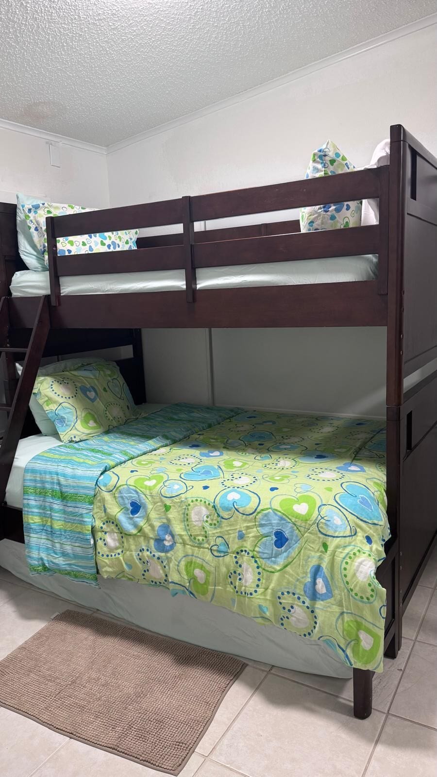 A bunk bed with a lower bunk, a green patterned bedspread, and a brown mat on the floor.