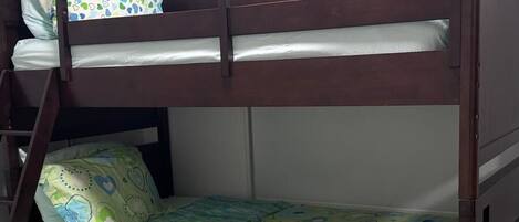 A bunk bed with a lower bunk, a green patterned bedspread, and a brown mat on the floor.