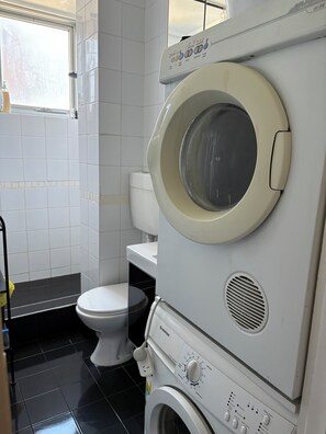 A compact laundry room with a stacked washer and dryer, a toilet, and a window. - Ocean Views Studio Apartment with Secure Parking, walk to shops and the beach. (St Kilda)