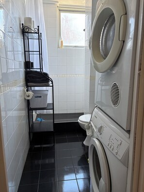 Ett litet och praktiskt tvättrum med tvättmaskin, torktumlare, en hylla med handdukar och ett fönster.