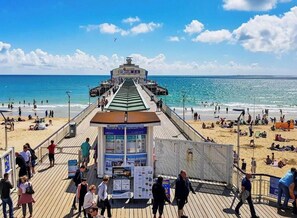 Um cais com uma bilheteria e placas de informação, que dá acesso a uma praia com pessoas e guarda-sóis.