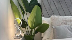 Um quarto com uma cama, cabeceira, uma planta na mesa de cabeceira e uma luminária ring light sobre uma prateleira.