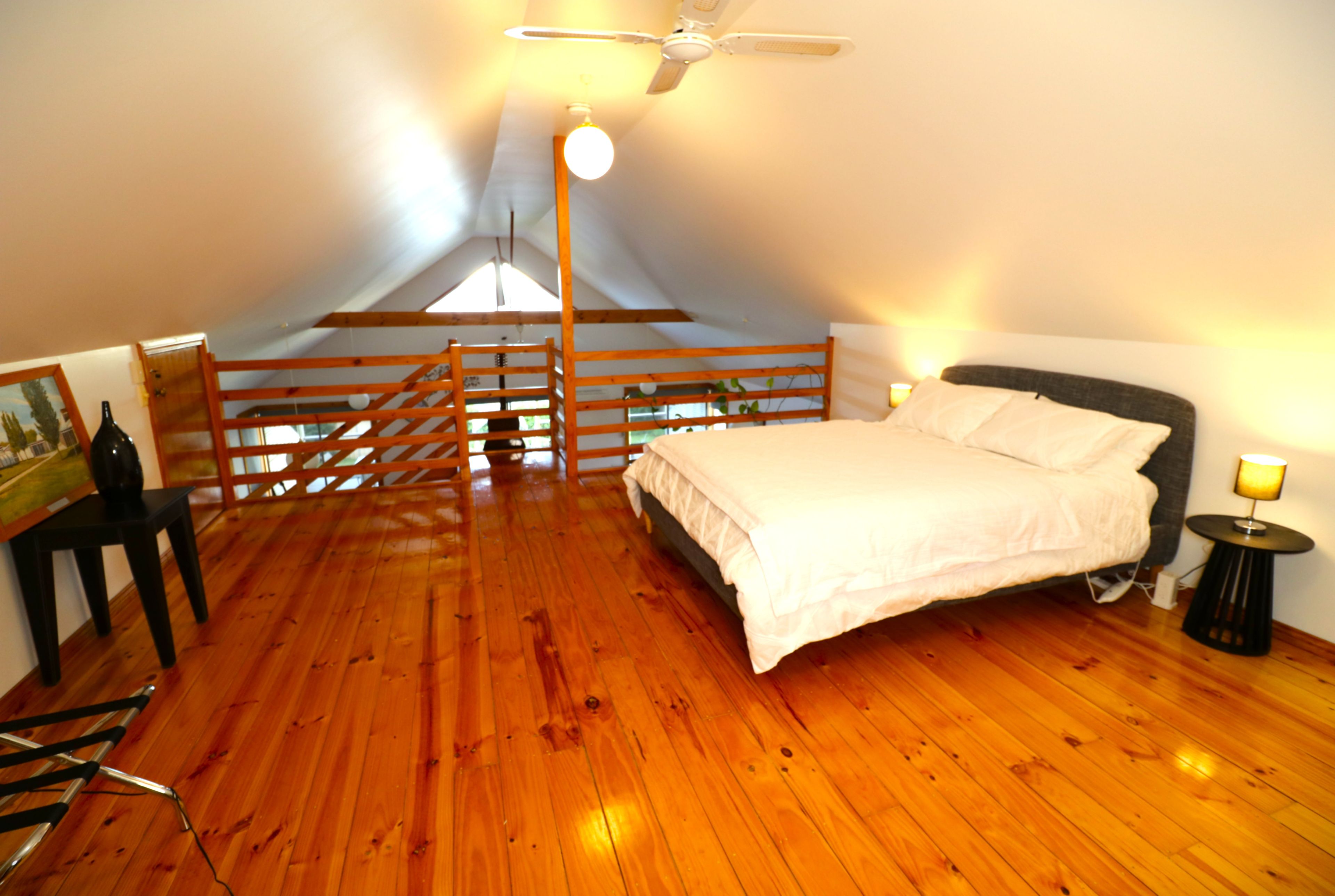 Une chambre avec un plancher en bois, un lit, une table de chevet, une lampe, un tableau et un escalier menant à un grenier.
