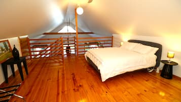 Une chambre avec un plancher en bois, un lit, une table de chevet, une lampe, un tableau et un escalier menant à un grenier.