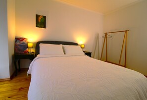 A neatly made bed with a white duvet and pillows, two bedside lamps, a painting on the wall, and a wooden floor.